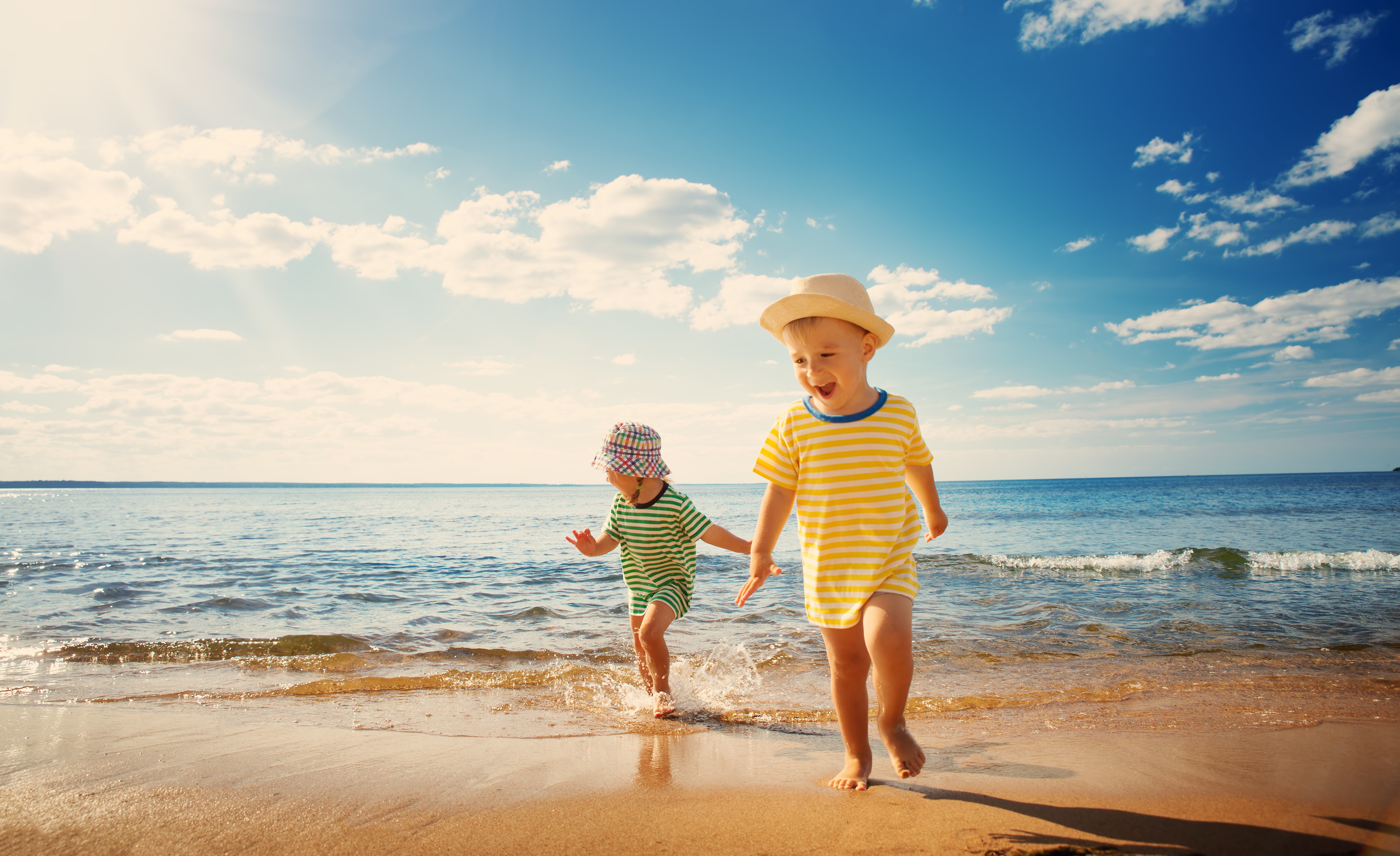 children running on the beach, playing in the water wearing sun hats